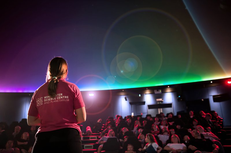 Woman with a ponytail and pink T-shirt looking out at an audience in a dome shaped planetarium